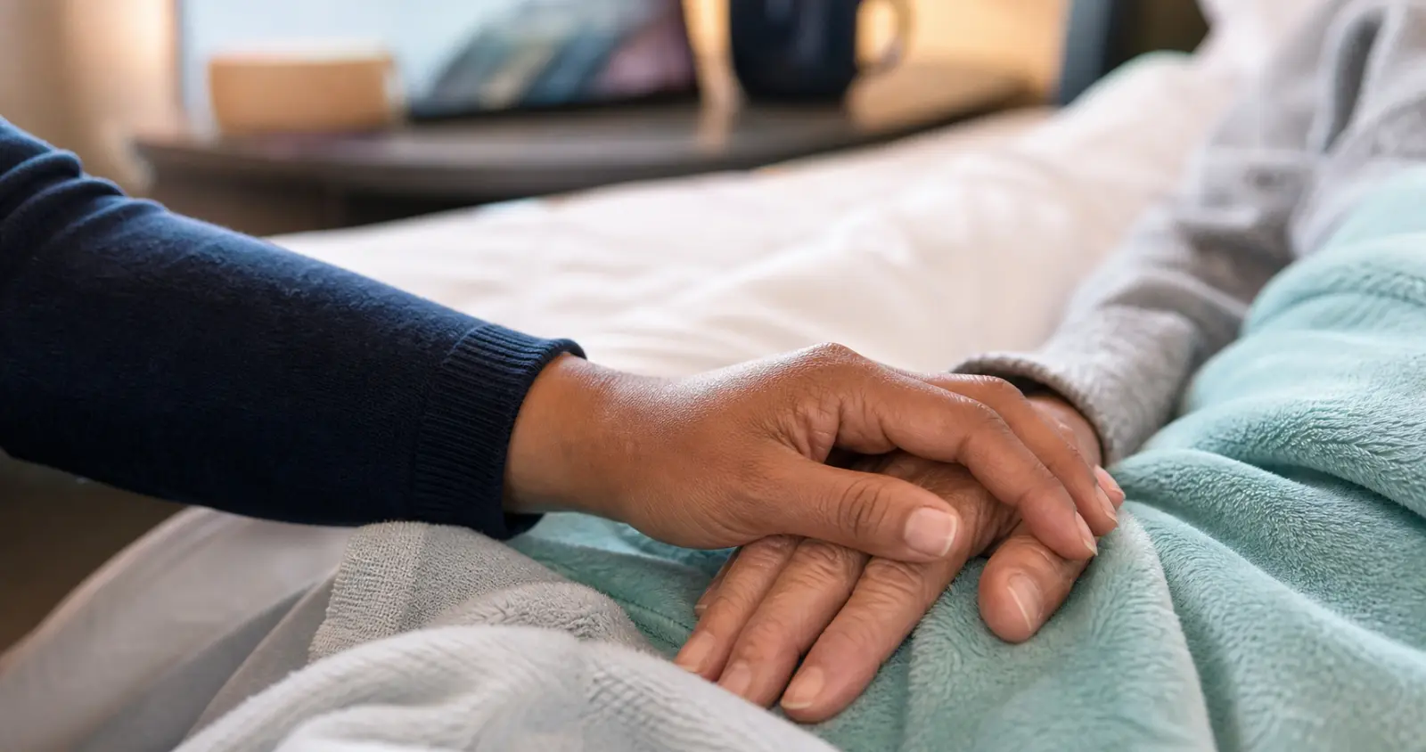 A close view of one person's hand resting gently over a patient's hand on a soft blanket: a quiet moment of reassurance.